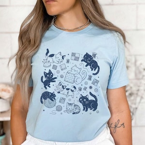 Peut inclure: T-shirt bleu clair orné d'un motif répétitif de chats espiègles, de bobines de fil et d'accessoires de couture. Les chats sont représentés dans diverses poses, jouant avec de la laine et interagissant avec des outils de couture. Le motif est bleu marine.