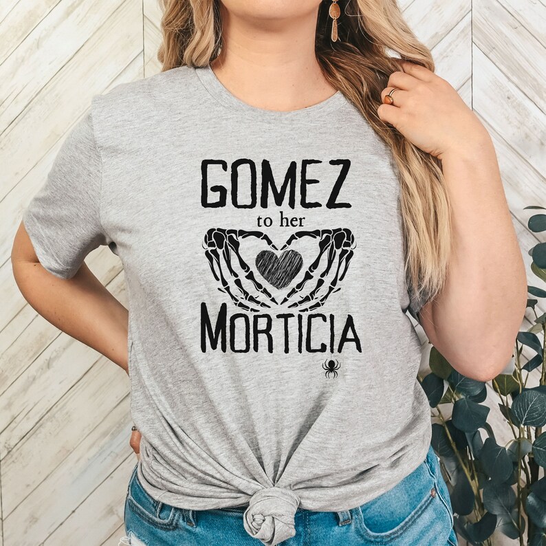 May include: A light grey t-shirt featuring the text "GOMEZ to her MORTICIA" in black. The design includes a heart held by skeleton hands and a small spider. The t-shirt is knotted at the waist.
