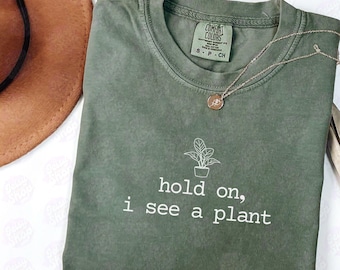 Peut inclure: T-shirt vert olive avec le texte blanc « hold on, i see a plant ». Un petit dessin de plante est au-dessus du texte. Un collier doré avec un pendentif rond est posé sur le t-shirt plié.