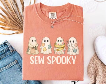 Könnte beinhalten: Terracottafarbenes T-Shirt mit dem Schriftzug "SEW SPOOKY" und vier Cartoon-Geisterfiguren, die jeweils Nähutensilien halten. Eine silberne Halskette mit Anhänger befindet sich auf dem Shirt. Das Shirt ist auf einer weißen Oberfläche gefaltet.