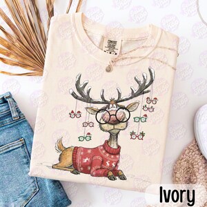 Könnte beinhalten: Elfenbeinfarbenes Sweatshirt mit einem Cartoon-Rentier, das eine Brille und einen roten Weihnachtspullover trägt. Das Rentier hat ein großes Geweih und ist von hängenden Brillen umgeben. Das Wort "Ivory" steht unten im Bild.