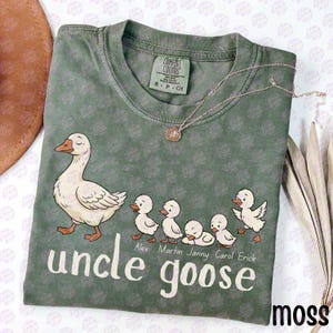 May include: A sage green short-sleeve t-shirt featuring a graphic of an adult goose followed by five small goslings. Below the goslings are the names Alex, Martin, Janny, Carol, and Erick. The text "uncle goose" is printed in large white letters.