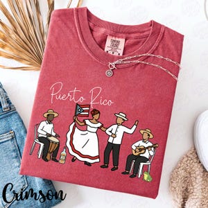Pu&ograve; includere: T-shirt color cremisi con la scritta "Puerto Rico" e una grafica di persone in abiti tradizionali. Il design include un batterista, una donna con una bandiera e due uomini, uno che suona la chitarra. Una collana d'argento &egrave; drappeggiata sulla maglietta.