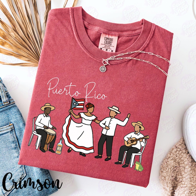 Pu&ograve; includere: T-shirt color cremisi con la scritta "Puerto Rico" e una grafica di persone in abiti tradizionali. Il design include un batterista, una donna con una bandiera e due uomini, uno che suona la chitarra. Una collana d'argento &egrave; drappeggiata sulla maglietta.