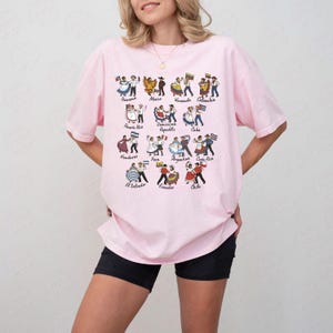 Puede incluir: Una camiseta rosa claro con un estampado colorido de figuras bailando que representan a varios pa&iacute;ses latinoamericanos. Los nombres de los pa&iacute;ses est&aacute;n impresos encima de las figuras. La camiseta es de corte holgado.