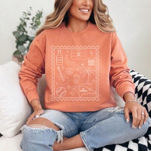 Pode incluir: Camisola coral com um desenho de contorno branco de s&iacute;mbolos da Rep&uacute;blica Dominicana dentro de uma borda de selo postal. O desenho inclui uma garrafa, banana, bandeira, tambor, guitarra, cafeteira, palmeira, basebol e mapa.