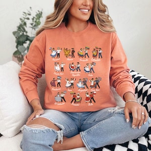 Puede incluir: Una sudadera de color coral con ilustraciones de parejas bailando, cada una representando un pa&iacute;s latinoamericano. Los nombres de los pa&iacute;ses est&aacute;n impresos encima de los bailarines. La sudadera tiene mangas largas y cuello redondo.