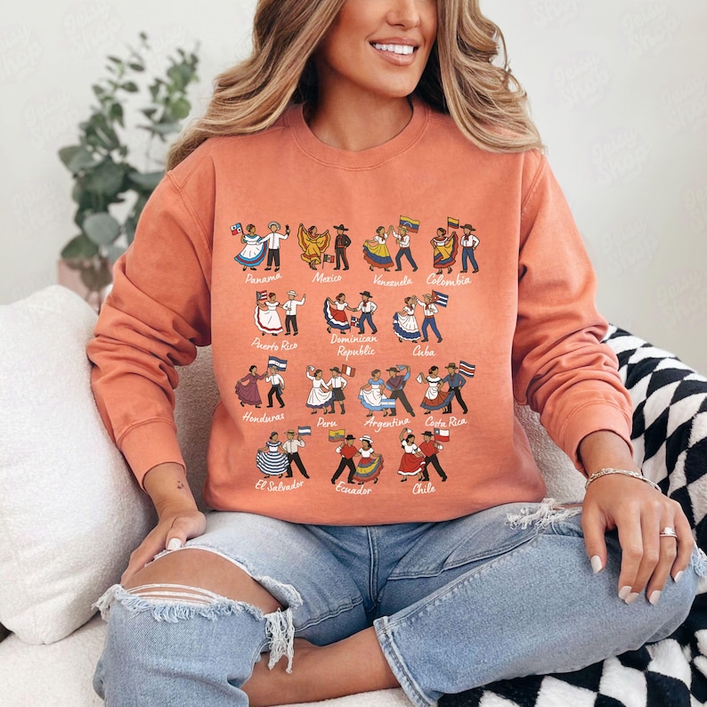 Puede incluir: Una sudadera de color coral con ilustraciones de parejas bailando, cada una representando un pa&iacute;s latinoamericano. Los nombres de los pa&iacute;ses est&aacute;n impresos encima de los bailarines. La sudadera tiene mangas largas y cuello redondo.