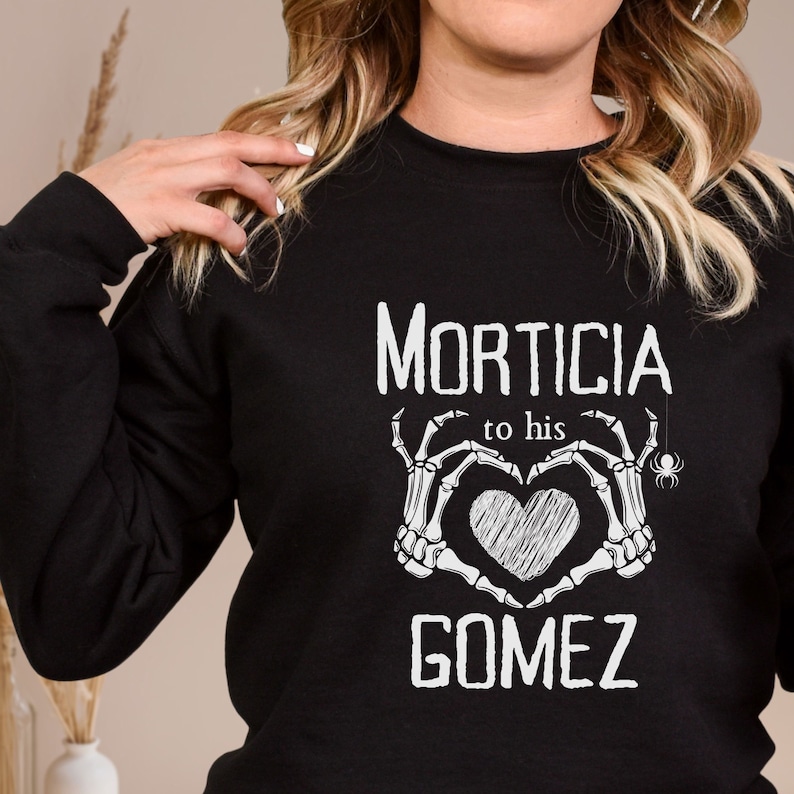 May include: Black sweatshirt with the words "MORTICIA to his GOMEZ" in white. The design features skeleton hands forming a heart around a sketched heart, with a spider hanging from the top.