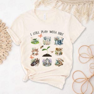 Puede incluir: Una camiseta beige con el texto "I Still Play With Dirt" impreso en la parte delantera. El texto está rodeado de una variedad de ilustraciones con temática de jardinería, que incluyen una carretilla, flores, herramientas de jardinería y una abeja.
