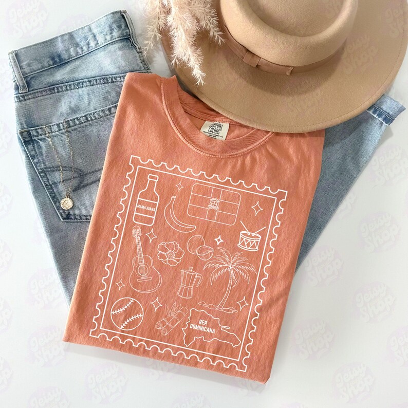 Pode incluir: Uma camiseta coral com um gr&aacute;fico branco de &iacute;cones da Rep&uacute;blica Dominicana, incluindo uma garrafa, banana, guitarra e palmeira. A camisa &eacute; combinada com jeans claros e um chap&eacute;u bege.