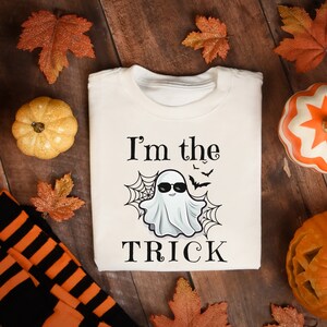 May include: A cream-colored crewneck sweatshirt featuring a graphic of a white ghost wearing black sunglasses, flanked by spiderwebs and small bats. The text "I'm the TRICK" is printed in black lettering above and below the ghost illustration.