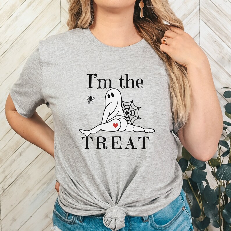 May include: A heather gray t-shirt featuring a black graphic of a ghost doing a split with a spider, a spiderweb, and a small red heart on its backside. The text "I'm the TREAT" is printed in black above and below the ghost illustration.