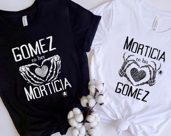 以下が含まれることがあります： 黒と白の2枚のTシャツ。それぞれ、骸骨の手で形作られたハートと、白と黒で印刷された「Gomez to her Morticia」と「Morticia to his Gomez」の文字が特徴です。