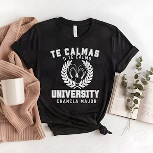 以下が含まれることがあります： 白い文字で「Te Calmas O Te Calmo University Chancla Major」と書かれた黒いTシャツ。テキストは、中央にビーチサンダルが置かれた葉の輪で囲まれています。