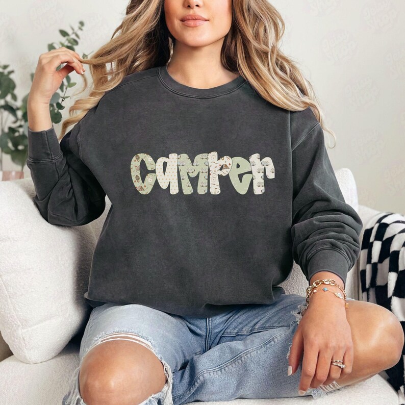 May include: Dark gray sweatshirt with the word "Camper" in a decorative font across the chest. The letters have a light green and beige pattern. The sweatshirt has long sleeves and a crew neck.
