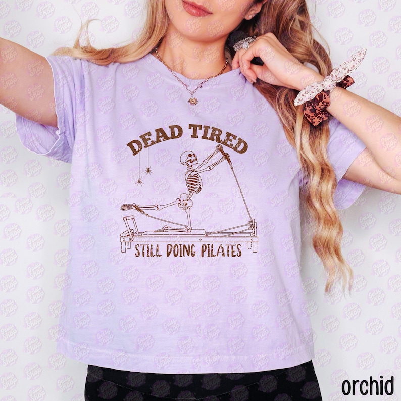 以下が含まれることがあります： ライトパープルのTシャツ。ピラティスのリフォーマーマシンで運動する骸骨の茶色のグラフィックが描かれています。骸骨の上には「DEAD TIRED」、下には「STILL DOING PILATES」という文字があり、クモの巣にぶら下がった2匹の小さなクモも描かれています。