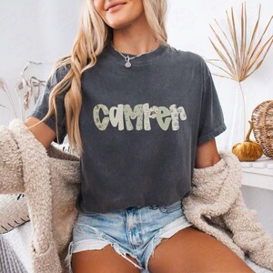 May include: Dark gray t-shirt with the word "Camper" in a green and tan camouflage pattern. The shirt is worn with denim shorts and a cream-colored cardigan. The overall theme is casual and outdoorsy.