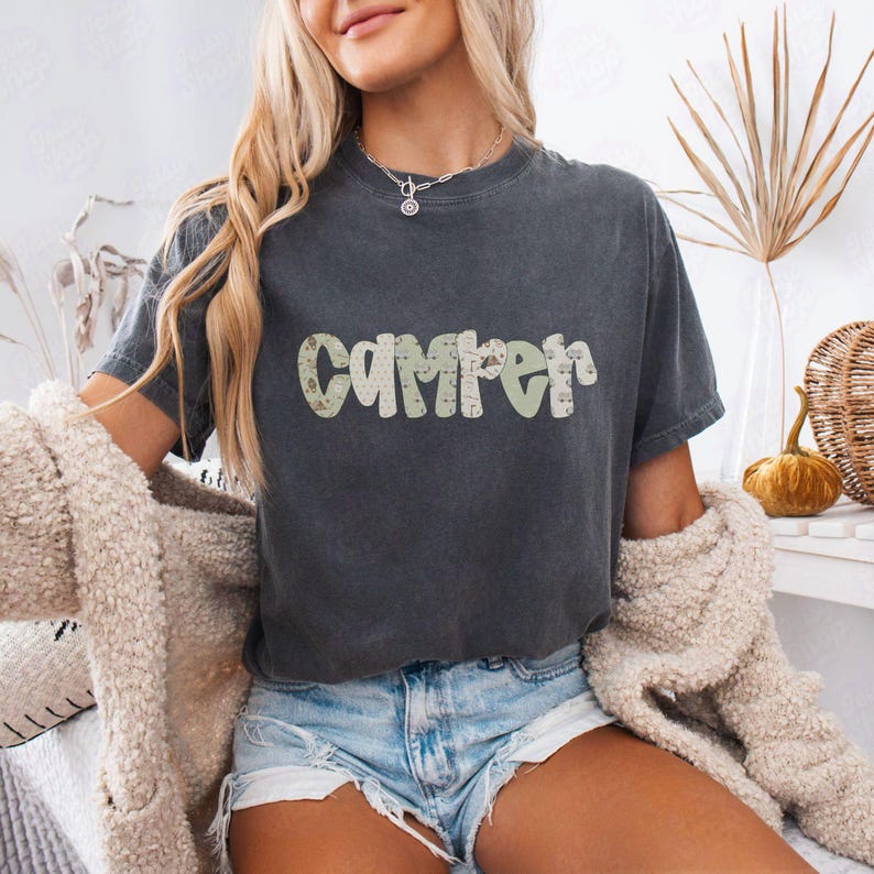 May include: Dark gray t-shirt with the word "Camper" in a green and tan camouflage pattern. The shirt is worn with denim shorts and a cream-colored cardigan. The overall theme is casual and outdoorsy.
