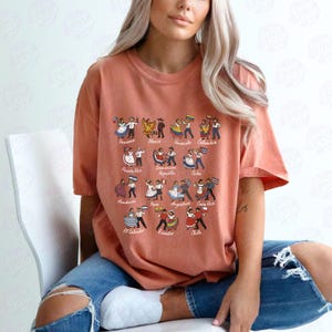 Puede incluir: Una camiseta de color coral con un gr&aacute;fico de figuras bailando que representan varios pa&iacute;ses latinoamericanos. Cada figura viste trajes tradicionales y sostiene una bandera. Los pa&iacute;ses est&aacute;n etiquetados con una fuente estilizada.