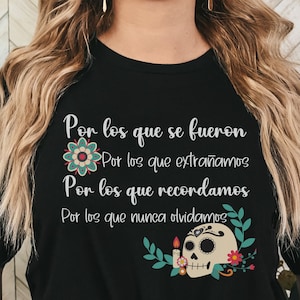 May include: Black t-shirt with white script and floral accents. The text reads "Por los que se fueron, Por los que extrañamos, Por los que recordamos, Por los que nunca olvidamos." Features a sugar skull and flowers.