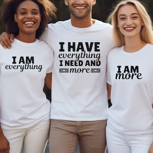 Op de afbeelding: Drie witte T-shirts met zwarte tekst. Het eerste shirt zegt "Ik ben alles". Het tweede shirt zegt "Ik heb alles wat ik nodig heb en meer". Het derde shirt zegt "Ik ben meer".