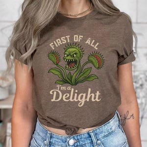 May include: Brown t-shirt featuring a green Venus flytrap graphic with the text "FIRST OF ALL I'M A Delight". The design includes a detailed illustration of the plant with open jaws and spiky leaves.