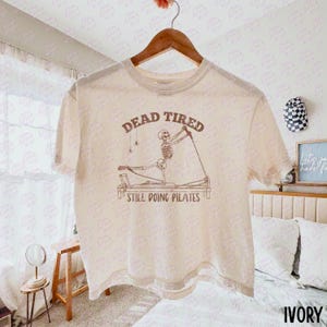 May include: An ivory short-sleeve t-shirt featuring a brown graphic of a skeleton on a pilates reformer machine. The text above the skeleton reads "DEAD TIRED" and the text below reads "STILL DOING PILATES." The shirt is displayed on a wooden hanger.