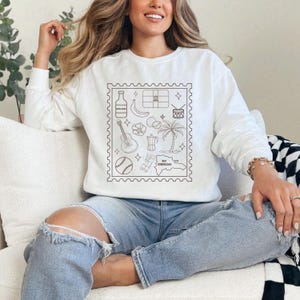 Pode incluir: Camisola branca de manga comprida com um desenho a preto de &iacute;cones da Rep&uacute;blica Dominicana dentro de uma borda de selo postal. O desenho inclui uma garrafa, uma banana, uma bandeira, um tambor, uma guitarra, uma flor, uma cafeteira, uma palmeira, uma bola de basebol e um mapa.