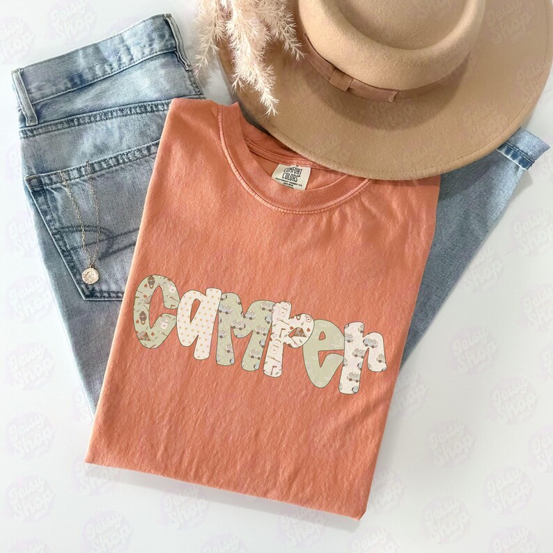 May include: A coral-colored t-shirt with the word "Camper" in a playful, patterned font. The shirt is laid on top of light-wash denim jeans and a tan fedora hat. A gold necklace is visible in the jeans pocket. Dried ornamental grass adds a decorative touch.