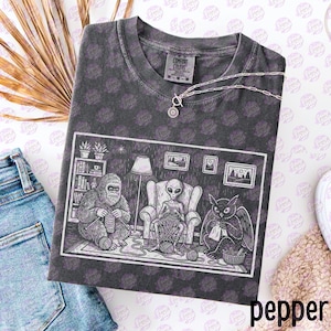 May include: Dark gray Comfort Colors t-shirt with a graphic of a Bigfoot, an alien, and an owl knitting. The shirt has a crew neck and a silver necklace. The word "pepper" is printed at the bottom.