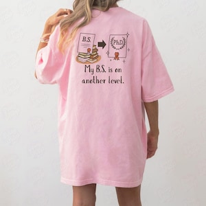 May include: A light pink oversized t-shirt with a graphic on the back. The graphic shows a progression from a B.S. degree to a Ph.D. degree, with the text "My B.S. is on another level."