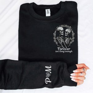 Puede incluir: Sudadera negra con un diseño de pareja de esqueletos dentro de una corona floral y el texto "Forever isn't long enough". La manga muestra las iniciales "P & M" en escritura blanca. El diseño está centrado en el pecho.
