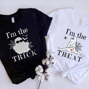 May include: A black t-shirt with white text reading I'm the TRICK and a ghost graphic, and a white t-shirt with black text reading I'm the TREAT and a ghost graphic with a red heart. Both shirts feature spiderweb designs and are displayed together.