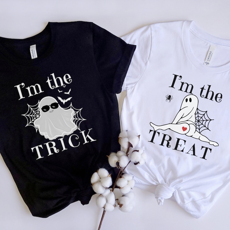 May include: A black t-shirt with white text reading I'm the TRICK and a ghost graphic, and a white t-shirt with black text reading I'm the TREAT and a ghost graphic with a red heart. Both shirts feature spiderweb designs and are displayed together.