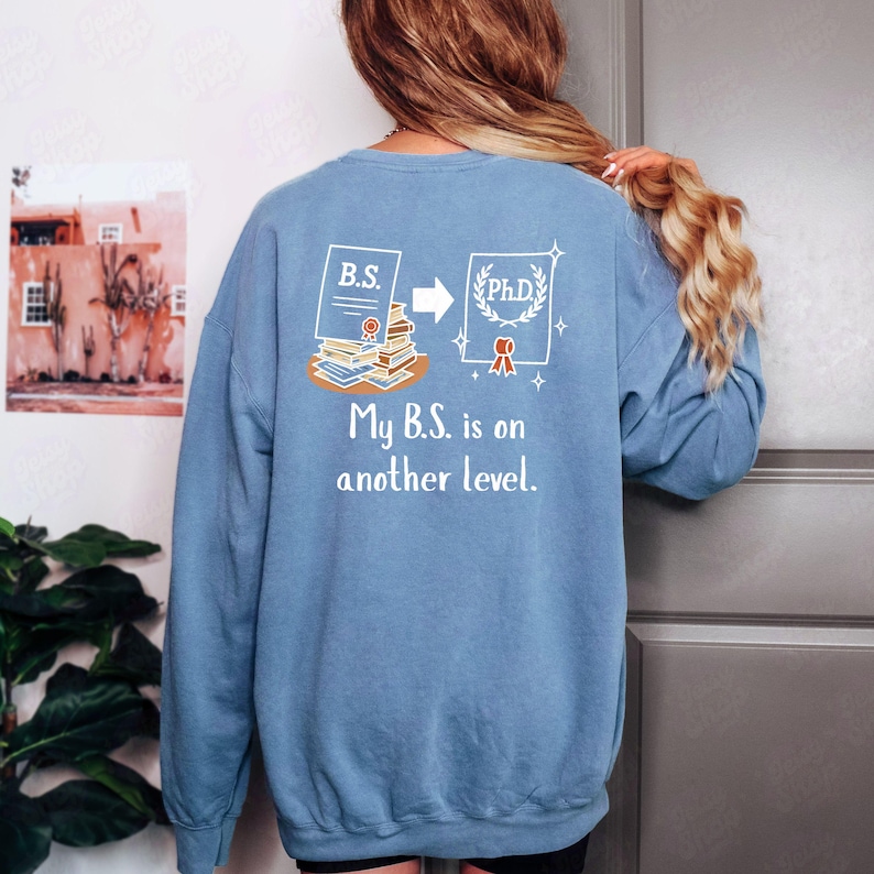 May include: A light blue sweatshirt with a graphic that shows a B.S. degree turning into a Ph.D. degree. The text "My B.S. is on another level" is below the graphic. The sweatshirt is a solid color.