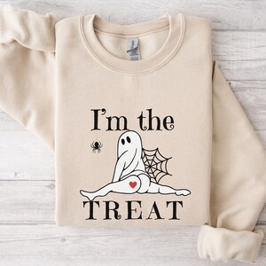 May include: A beige crewneck sweatshirt featuring a graphic of a ghost doing a split with a spider web behind it, a small spider hanging from a thread, and a red heart on its rear. The text reads I'm the TREAT in black, stylized lettering.