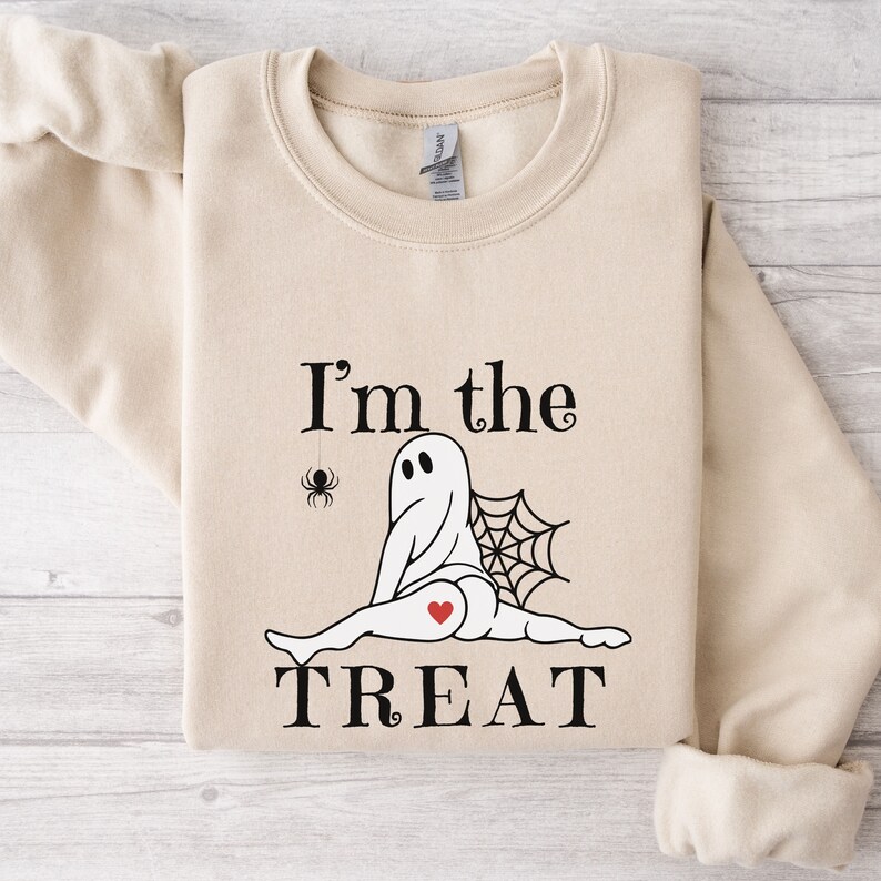 May include: A beige crewneck sweatshirt featuring a graphic of a ghost doing a split with a spider web behind it, a small spider hanging from a thread, and a red heart on its rear. The text reads I'm the TREAT in black, stylized lettering.