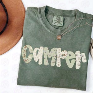 May include: Olive green t-shirt with the word "Camper" in a bubble font. The letters are decorated with camping-themed patterns. A silver necklace with a pendant is draped across the shirt.