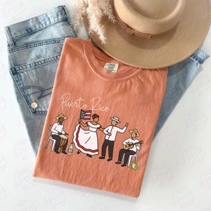 Pu&ograve; includere: Una maglietta color corallo con la scritta "Puerto Rico" e una grafica di persone che ballano e suonano strumenti. La maglietta &egrave; abbinata a jeans azzurri e un cappello color cammello.
