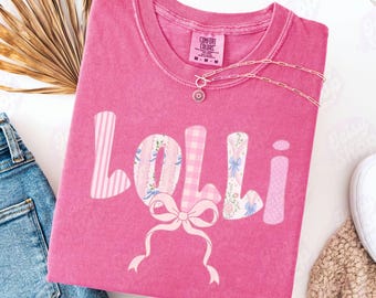 以下が含まれることがあります： ピンク色のTシャツ。名前「Lolli」が遊び心のあるフォントで書かれています。文字はストライプ、ギンガムチェック、花柄、水玉模様で装飾されています。名前の下にはピンクのリボンがあります。太陽のペンダントが付いたシルバーのネックレスがシャツの上にあります。