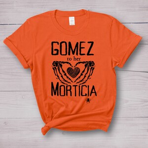 May include: An orange t-shirt featuring the words "GOMEZ to her MORTICIA" in black. The design includes a heart formed by skeleton hands and a small spider graphic. The shirt is made of a soft material.