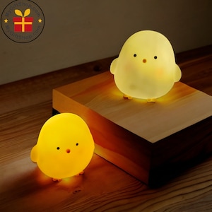 May include: Two yellow chick-shaped night lights. The lights are glowing, with black eyes and orange feet. One light is on a wooden block, and the other is on a wooden surface. The lights are designed to look like cartoon chicks.