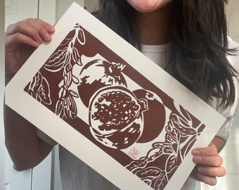 May include: A print featuring a pomegranate design in shades of brown and white. The artwork showcases a detailed illustration of pomegranates, with one cut open to reveal the seeds. The print has a rectangular shape.