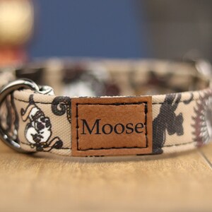 Op de afbeelding: Een beige hondenhalsband met een zwart-wit paisley-patroon en een bruin leren label met de tekst "Moose".