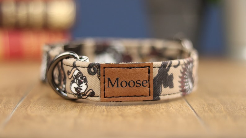 Op de afbeelding: Een beige hondenhalsband met een zwart-wit paisley-patroon en een bruin leren label met de tekst "Moose".