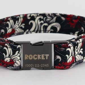 Puede incluir: Collar para perro de patrón negro y rojo con una hebilla plateada. La hebilla tiene la palabra "ROCKET" y un número de teléfono grabado.
