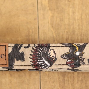 Op de afbeelding: Een beige hondenhalsband met een bruin leren patch waarop "Moose" staat en een kleurrijke tattoo-ge&iuml;nspireerde print met een zwarte panter, een Indiaanse hoofdtooi, een adelaar en een roos.