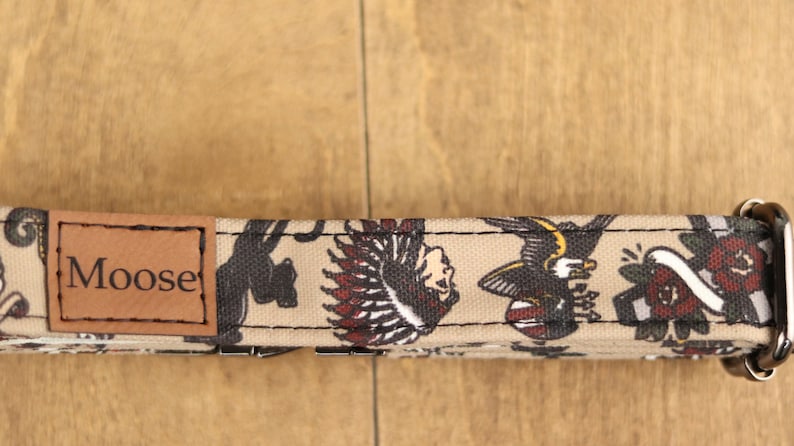 Op de afbeelding: Een beige hondenhalsband met een bruin leren patch waarop "Moose" staat en een kleurrijke tattoo-ge&iuml;nspireerde print met een zwarte panter, een Indiaanse hoofdtooi, een adelaar en een roos.