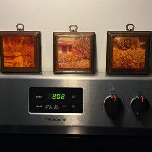 May include: Three square framed pictures with landscape scenes, each with a dark wood frame and a small metal loop at the top for hanging. The frames are resting on a stainless steel Frigidaire oven. The oven display shows the time as 8:08.